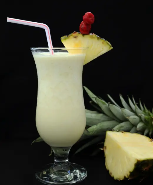 Creamy piña colada cocktail served in a hurricane glass with a pineapple slice and cherry garnish on a black background.